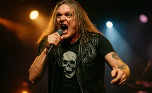 A male heavy metal singer with long blonde hair shouts into a microphone while wearing a black t-shirt with a skull graphic and a leather vest.