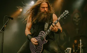 A long-haired, bearded guitarist plays a purple and black spiral electric guitar on stage under bright lights.