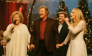 A group of people singing on stage with Christmas trees in the background.