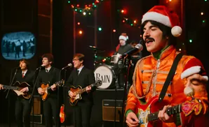 A band in Santa hats performs in front of Christmas lights. One guitarist wears an orange jacket.
