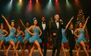 A male singer in a tuxedo performs with a chorus line of female dancers in sparkling blue outfits on a stage with Christmas trees.