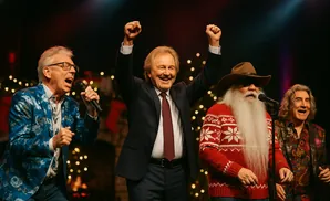 Four men singing on stage, one with a long white beard, festive lights in the background.