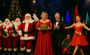 Christmas performers on stage: Santas, singers in red dresses, a man in a tuxedo, and an elf.