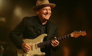 A man in a hat and suit plays a light-colored electric guitar.