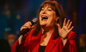 Singer in a red blazer holds a microphone, smiling and raising her hand with a musical note earring.