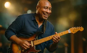 A smiling bald man plays a sunburst electric guitar on a stage.