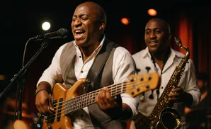 A bald Black man sings and plays a bass guitar on stage with a saxophonist in the background.