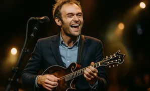 A man in a suit laughs while playing a mandolin on stage.