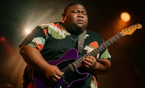 A Black man plays a purple electric guitar on stage with lights in the background.
