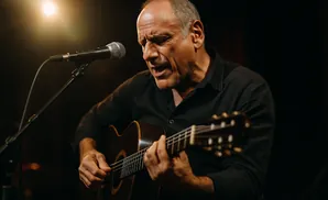 A man sings into a microphone while playing an acoustic guitar on stage.
