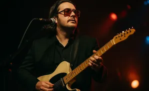 A man in a black suit and sunglasses plays a light-colored electric guitar on a dark stage with red and blue lights.