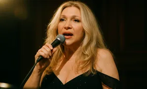 Woman with long blonde hair singing into a microphone on a dimly lit stage.