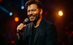 A smiling man in a dark suit holds a microphone on stage with blurred lights behind him.