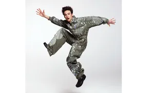 A person in a paint-splattered jumpsuit jumps with arms outstretched against a white background.
