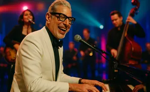 Jeff Goldblum plays piano in a white suit, laughing, with a band performing behind him.