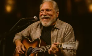 A smiling older man with a grey beard plays an acoustic guitar and sings into a microphone.