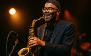 A Black man wearing glasses and a skullcap plays a saxophone on stage, smiling under a spotlight.