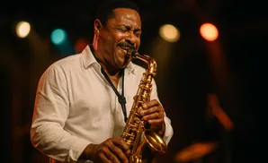 A Black man in a white shirt plays a saxophone with his eyes closed in front of blurry colorful lights.