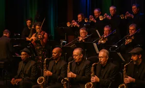 A jazz orchestra performs on stage, with musicians playing brass instruments and a violinist.