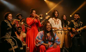 A diverse band plays music together on a dimly lit stage, featuring brass instruments, drums, and a bass guitar.