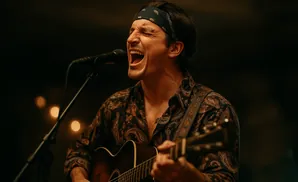 Man singing and playing guitar with eyes closed, wearing a bandana and paisley shirt.
