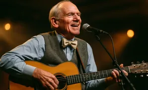 A smiling older man with gray hair plays an acoustic guitar and sings into a microphone.