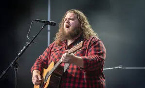 A man with a long beard and curly hair sings into a microphone while playing an acoustic guitar on stage.