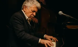 A man with gray hair plays piano with his eyes closed, wearing a suit and tie.