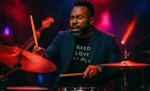 A Black male drummer plays with his eyes closed, holding drumsticks, with red and purple stage lighting.