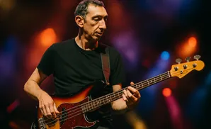 A man in a black t-shirt plays a bass guitar on a dimly lit stage with colorful lights in the background.