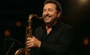 A smiling man plays a saxophone, with a microphone visible in the background.