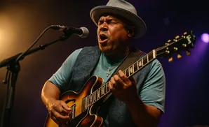 Man in a fedora sings and plays a hollow-body electric guitar on stage.