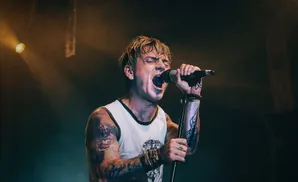 A tattooed man singing intensely into a microphone on a dark stage with a spotlight.