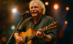 A man with grey hair and a mustache plays an acoustic guitar and sings into a microphone on stage.