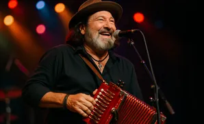 A man in a hat and black shirt plays a red accordion, smiling into a microphone on a stage with colorful lights.