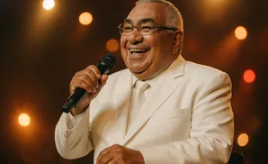 A man in a white suit and tie, wearing glasses, smiles while holding a microphone.