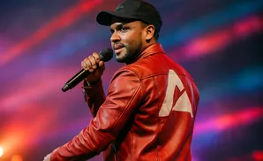 Man in a red leather jacket and black cap holding a microphone, performing on stage with colorful lights.