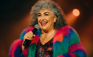 A smiling woman with grey curly hair wears a colorful fur coat and sequined top while holding a microphone.
