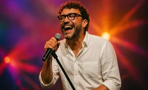 A man with curly hair and glasses sings into a microphone on a colorful stage.