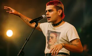 A musician with red and blonde hair sings into a microphone, wearing a t-shirt with a portrait.