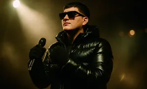 Man in shiny black puffer jacket and sunglasses holds a microphone while illuminated by a spotlight.