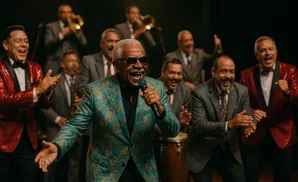 A band performs, with a lead singer in a teal sequined jacket holding a microphone, backed by musicians in suits and sparkly red jackets.