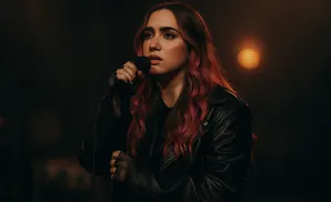 A young woman with long, wavy pink hair sings into a microphone while wearing a black leather jacket and fishnet gloves.