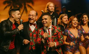 Band members perform on stage, with two men in floral suits, one in a leather jacket, and two women in sparkly outfits.