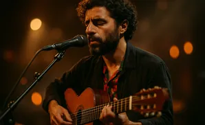 A man with curly hair and a beard sings into a microphone while playing an acoustic guitar.