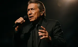 A man in a black suit and tie sings into a microphone, his face lit from the side.