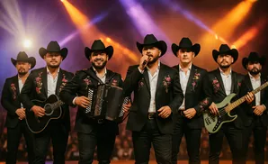 A band of six men wearing cowboy hats and black suits with floral embroidery, performing on stage.