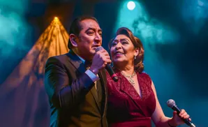 A man and woman sing into microphones on a dimly lit stage with blue and orange spotlights.