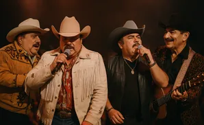 Four men in cowboy hats perform on stage, two singing into microphones, one playing guitar.