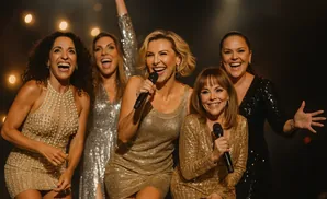 Five women in sparkly dresses singing into microphones on a stage.